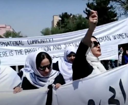 A group of Afghan women march in the street holding a large sign.